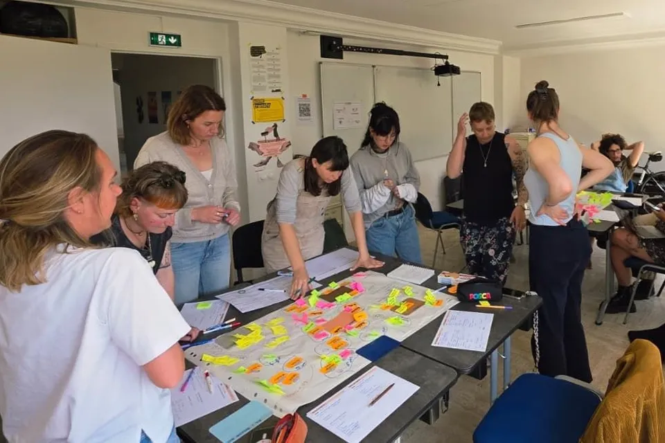 Atelier du collectif : debout autour de tables mises bout à bout, plusieurs personnes travaillent sur un grand plan couvert de post-it de couleur