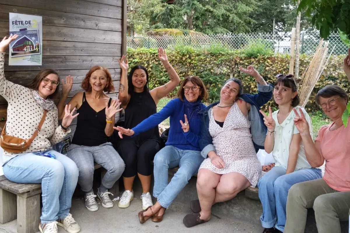 Sept bénévoles du collectif, assises ensemble devant un bâtiment en bois, sourient et lèvent la main en signe de victoire