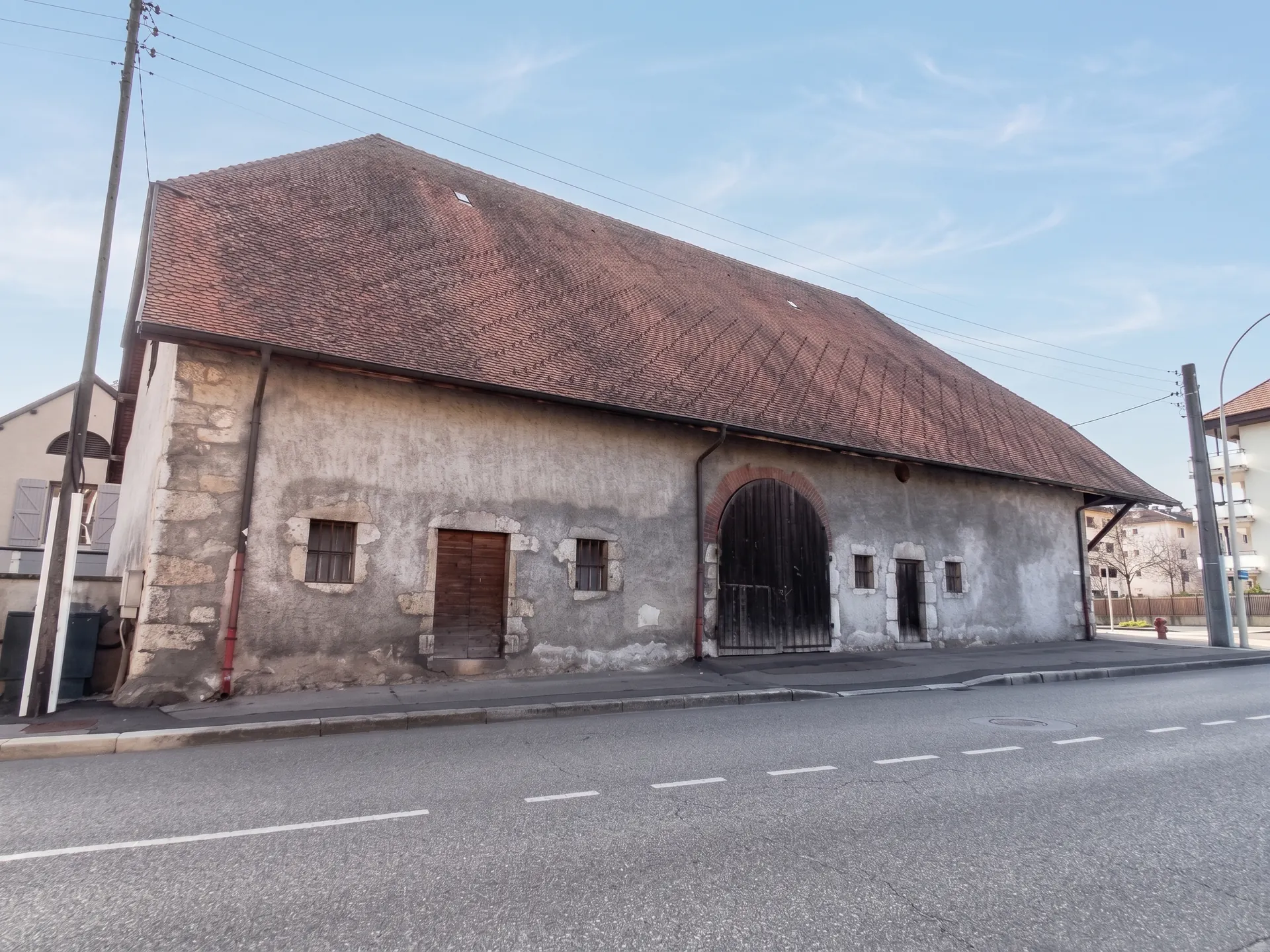 La ferme depuis la rue&nbsp;: un long mur de pierre enduit percé d'une porte charretière en arc, sous un grand toit de tuiles