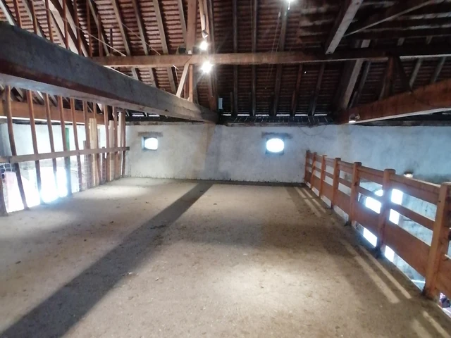 L'étage de la grange&nbsp;: un sol de béton, une barrière de bois le long du vide et la charpente au-dessus, éclairée par deux oculus