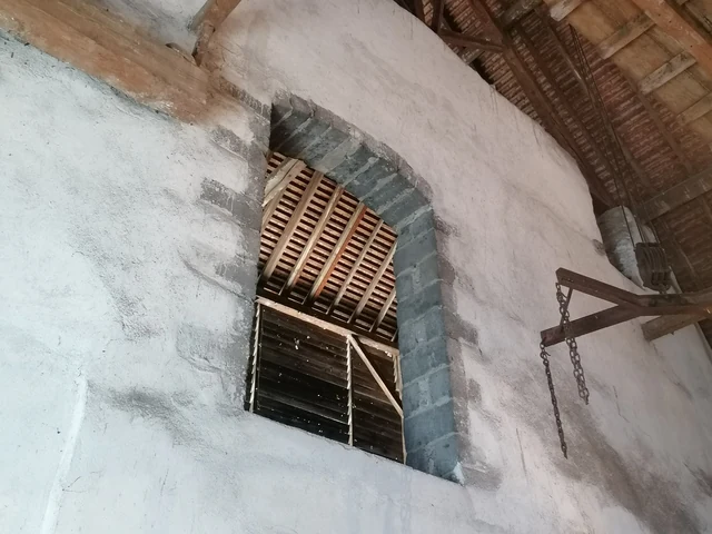Un mur blanchi percé d'une ouverture encadrée de blocs gris et fermée par des planches, sous la charpente