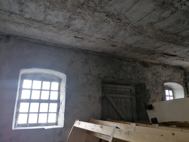 Une pièce sombre sous un plafond de béton&nbsp;: une fenêtre à petits carreaux, une vieille porte de bois et des planches entassées